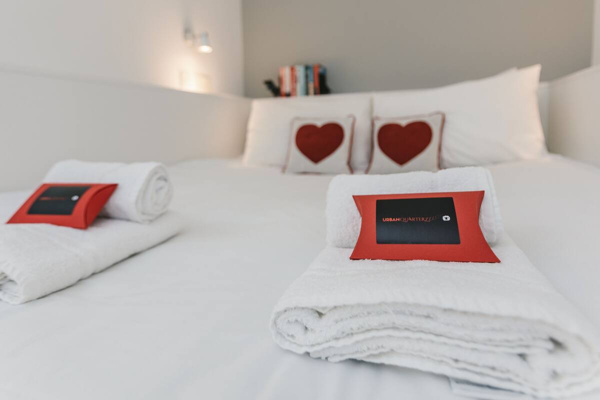 A neatly made bed with white linens and red heart-shaped pillows, a folded towel with a red tag, and a bedside table with a red device.