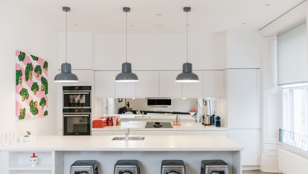 A modern kitchen with white cabinetry, a central island with bar stools, pendant lighting, and a large window providing natural light. - Urban Quarters Serviced Apartments (Dundee)