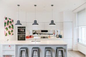 A modern kitchen with white cabinetry, a central island with bar stools, pendant lighting, and a large window providing natural light. - Urban Quarters Serviced Apartments (Dundee)
