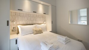 A neatly made bed with white linens and towels, a headboard with a textured design, and a wall-mounted mirror. - Urban Quarters Serviced Apartments (Dundee)