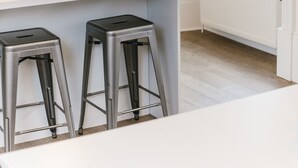 A modern kitchen with a white countertop, two bar stools, a hanging light fixture, and a window with a view of the city. - Urban Quarters Serviced Apartments (Dundee)