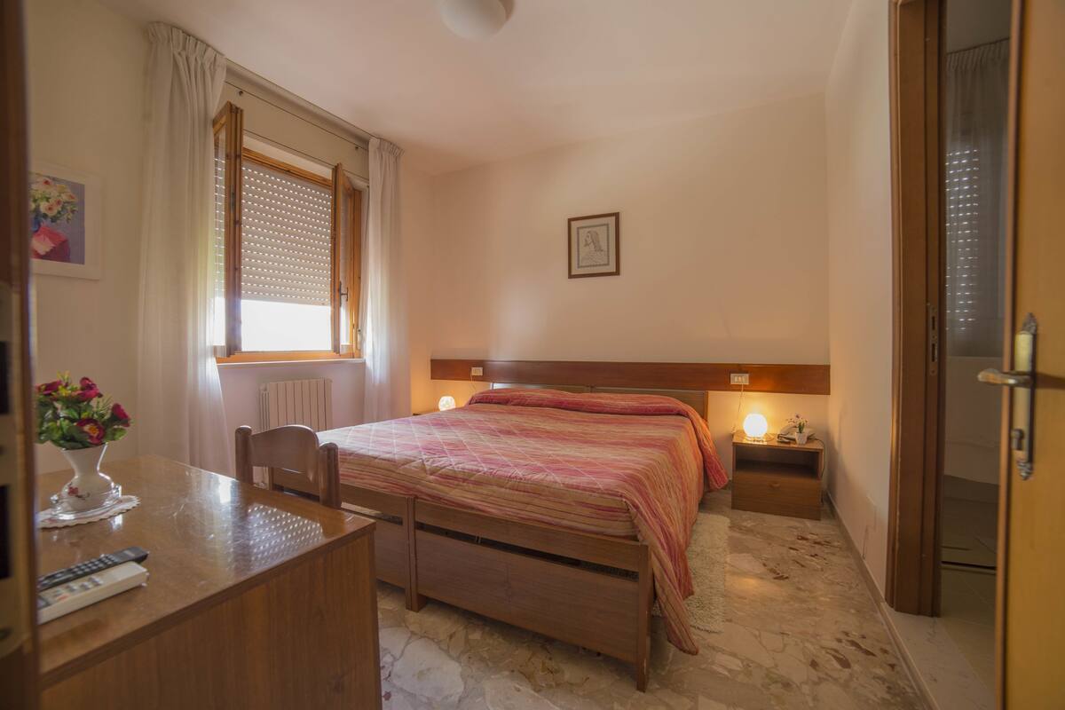Een slaapkamer met een houten bed, een nachtkastje met een lamp, een raam met jaloezieën en een kleine tafel met een bloevas.