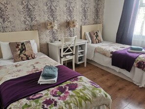Two single beds with floral-patterned bedding and matching pillows, a small desk with a lamp, and elegant wall decor create a comfortable and inviting space.
