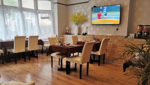 A dining area with wooden flooring, beige chairs, and a table set for four. A television mounted on the wall displays a tennis match. - Adams Guest House (Oxford)