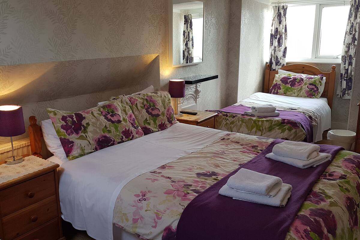 Une chambre d’hôtel avec deux lits, une literie à motifs floraux, une table de chevet avec une lampe et une fenêtre avec des rideaux.