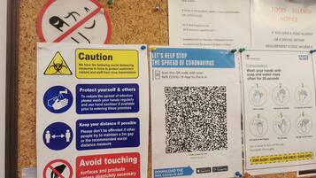 A bulletin board with various COVID-19 safety notices, including social distancing measures, hand washing instructions, and a QR code for the NHS COVID-19 app.