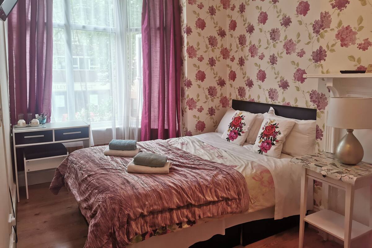 Un lit bien fait, agrémenté d’oreillers à motifs floraux et d’une couvre-lit rose, une table de chevet avec une lampe et une fenêtre ornée de rideaux légers.