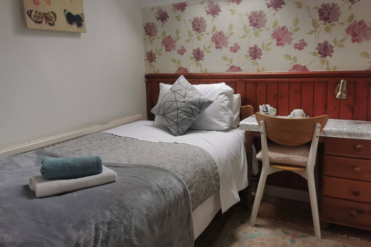 Une chambre à coucher comprenant un lit, un bureau, une chaise et un mur recouvert d’un papier peint à motifs floraux.