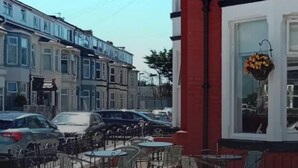 Vista callejera con coches aparcados, una fila de casas adosadas y terraza con mesas y sillas.