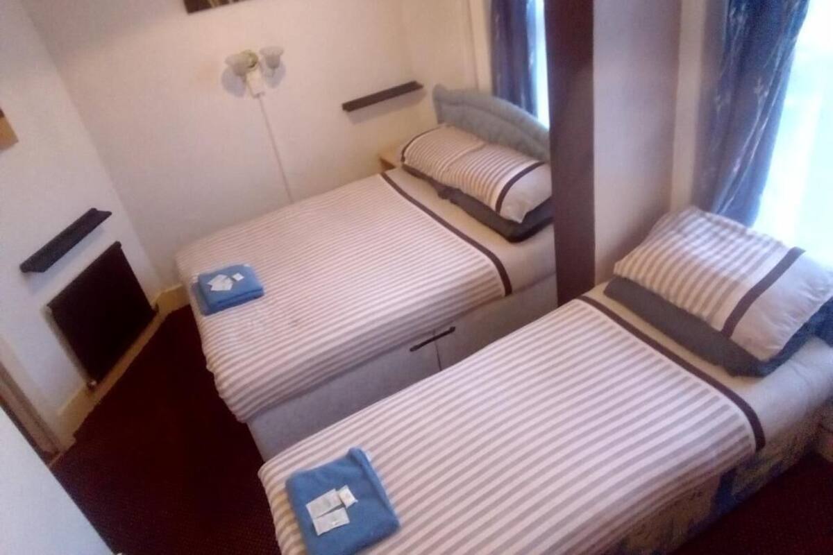 A small, compact room with two single beds, a bedside table with a lamp, a power outlet, and a window with curtains.