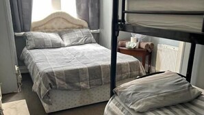 Un dormitorio con dos camas, una con cabecera y otra litera. Hay una mesita con una botella y una caja de pañuelos.