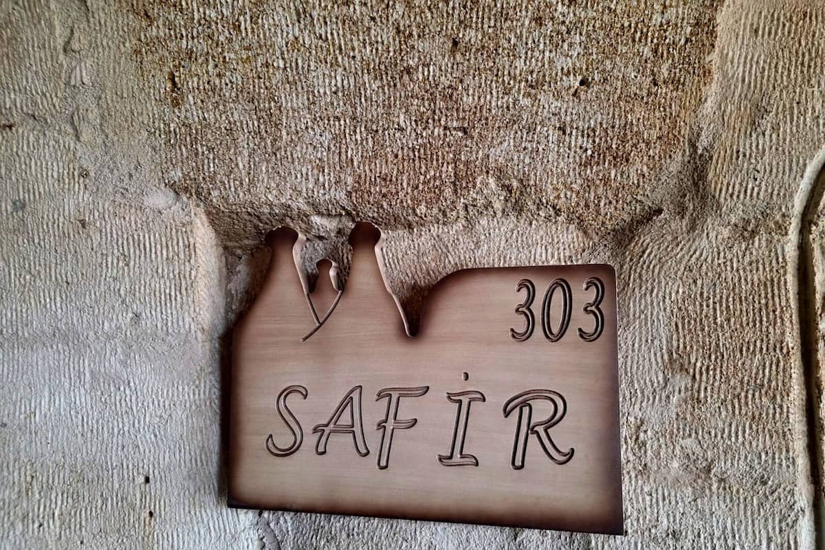 Un senyal de fusta amb el número 303 i la paraula "SAFIR" gravada, col·locat davant d'una paret de pedra amb textura.
