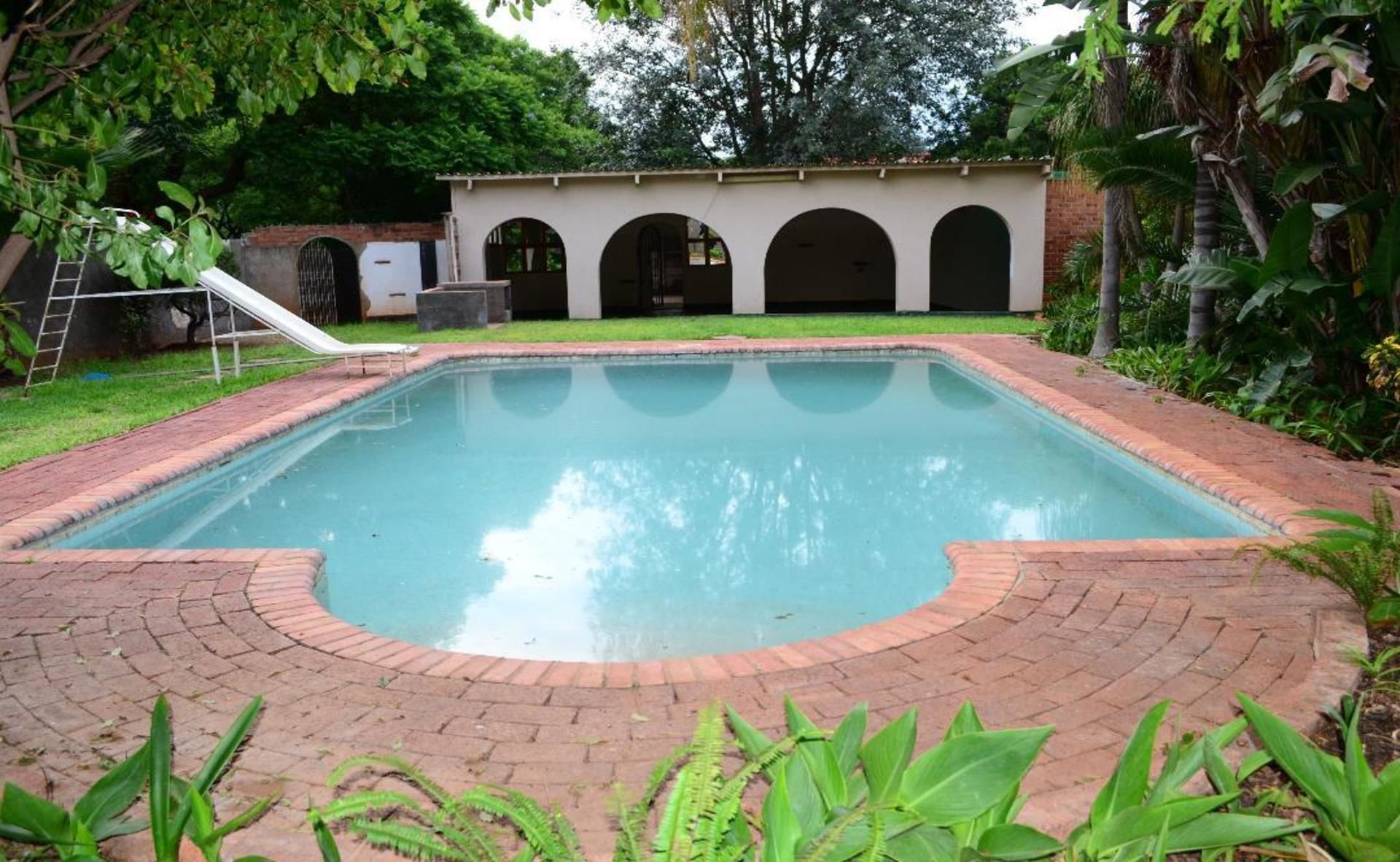 A rectangular swimming pool with a brick border, surrounded by a grassy area and a building with arched doorways.