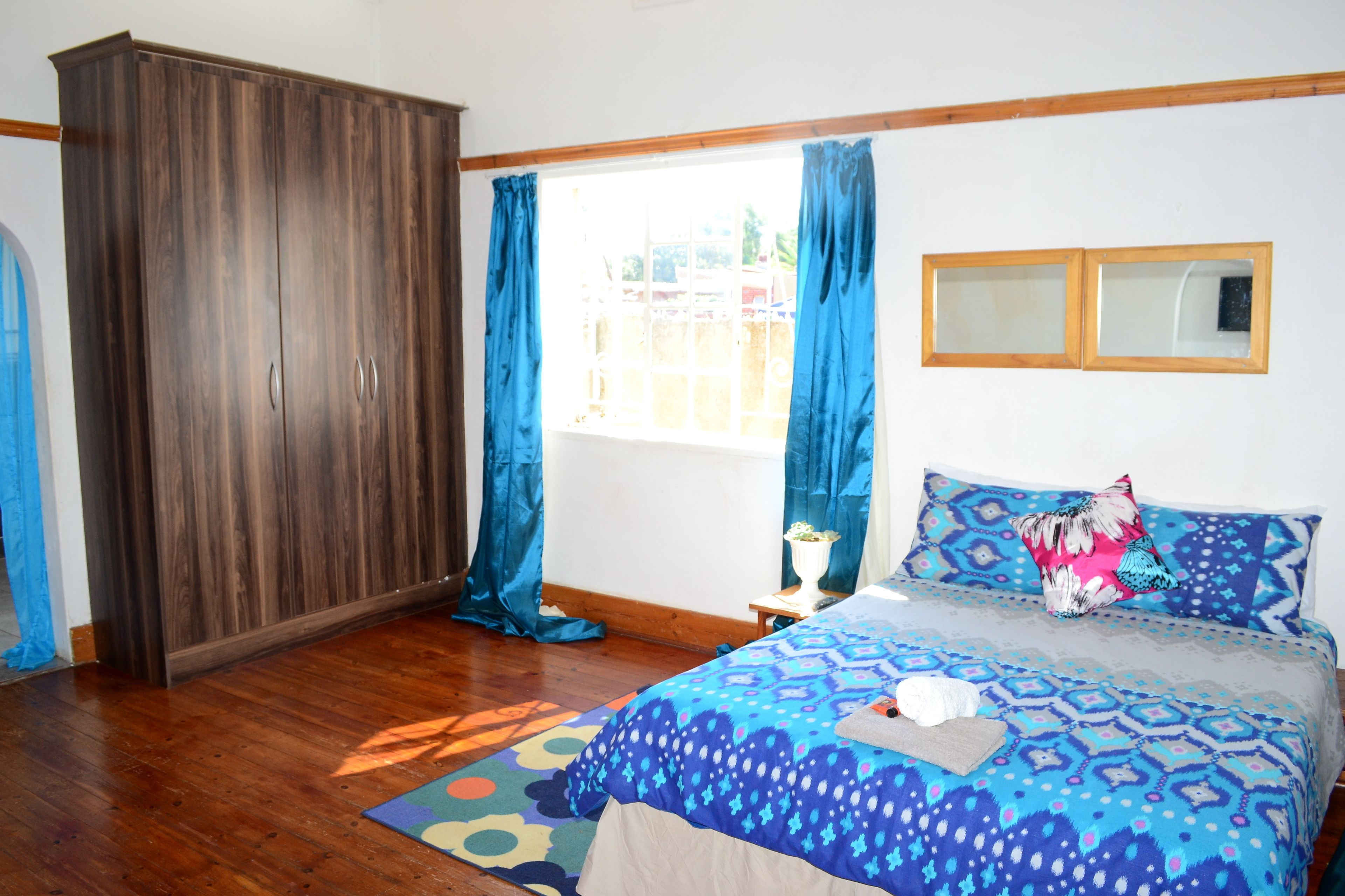 Un dormitorio con cama, armario, ventana con cortinas y una mesita con una planta.