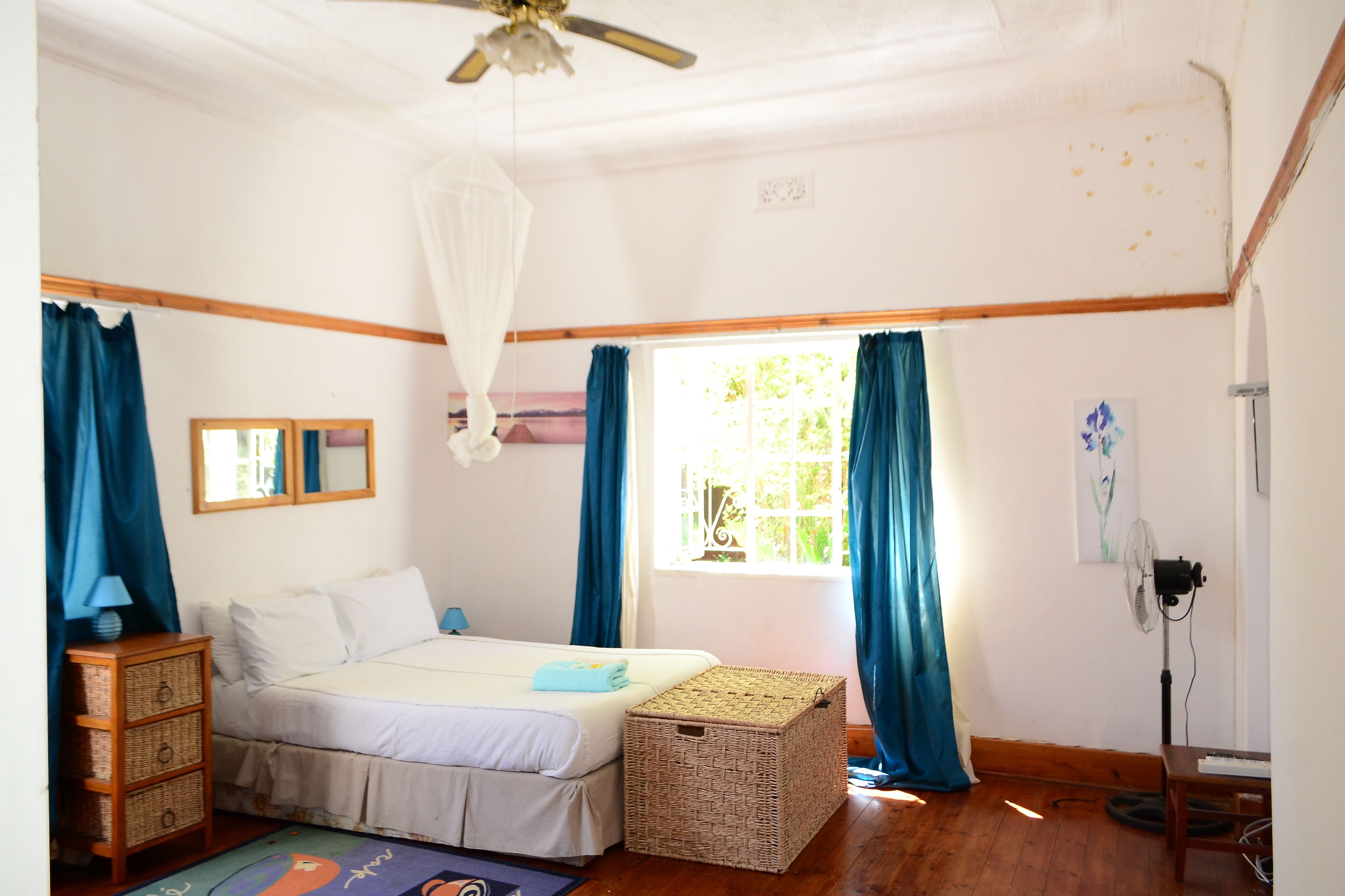 A bedroom with a bed, a bedside table, a standing fan, a window with curtains, and a ceiling fan.