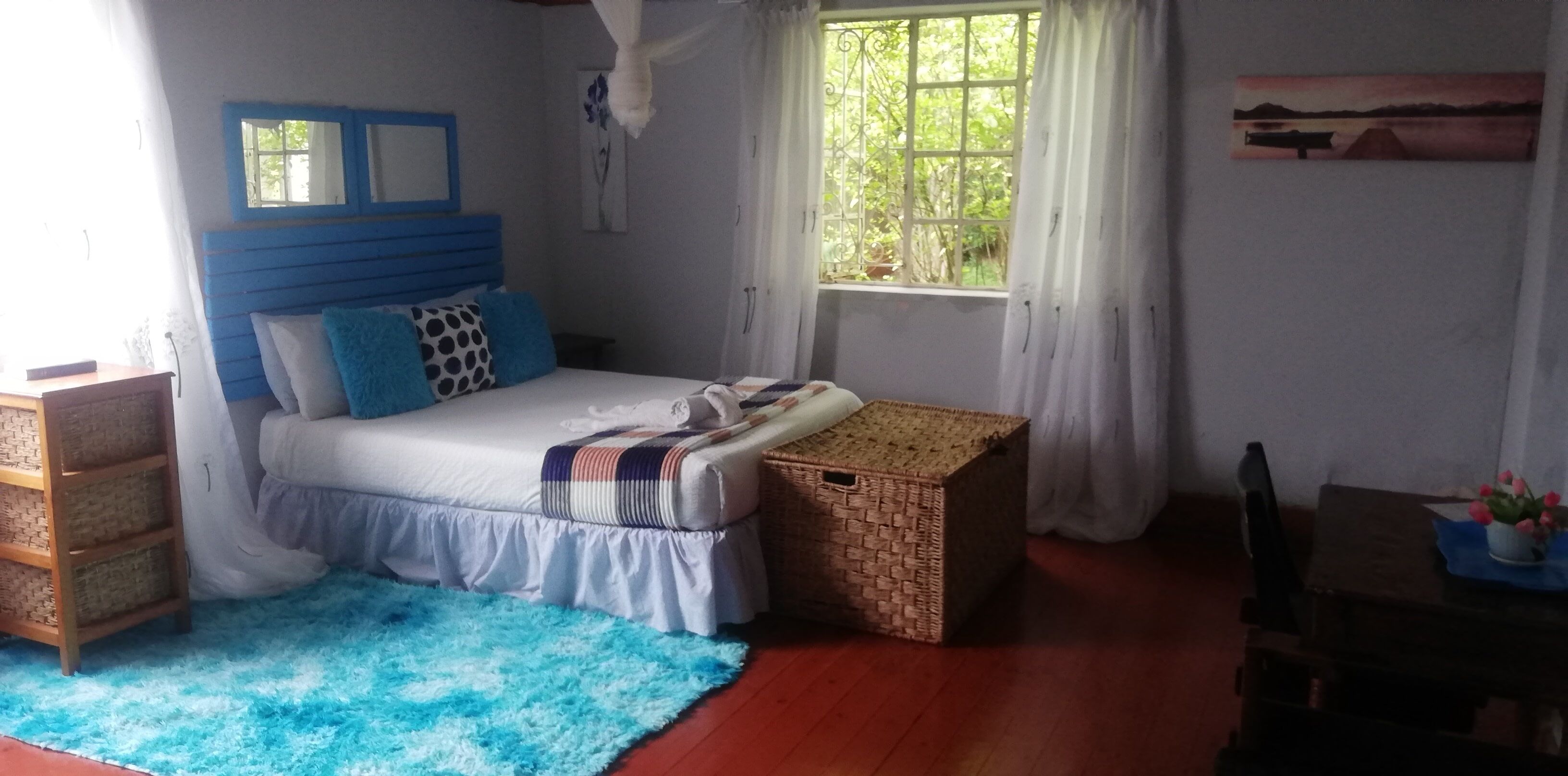 Une chambre à coucher avec un lit, une fenêtre avec des rideaux, une table de chevet ornée d’un vase de fleurs et un tableau accroché au mur.