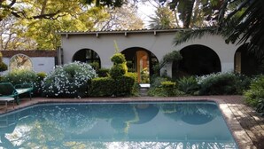 A swimming pool surrounded by lush greenery and a building with arched doorways. - Breeze Guest House (Bulawayo)