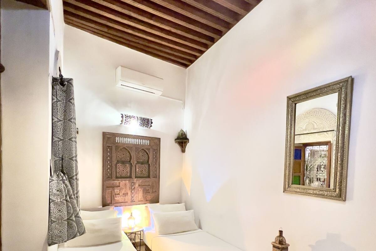 A room with two beds, a patterned curtain, a decorative wall panel, a small table with a lamp, and a mirror on the wall.