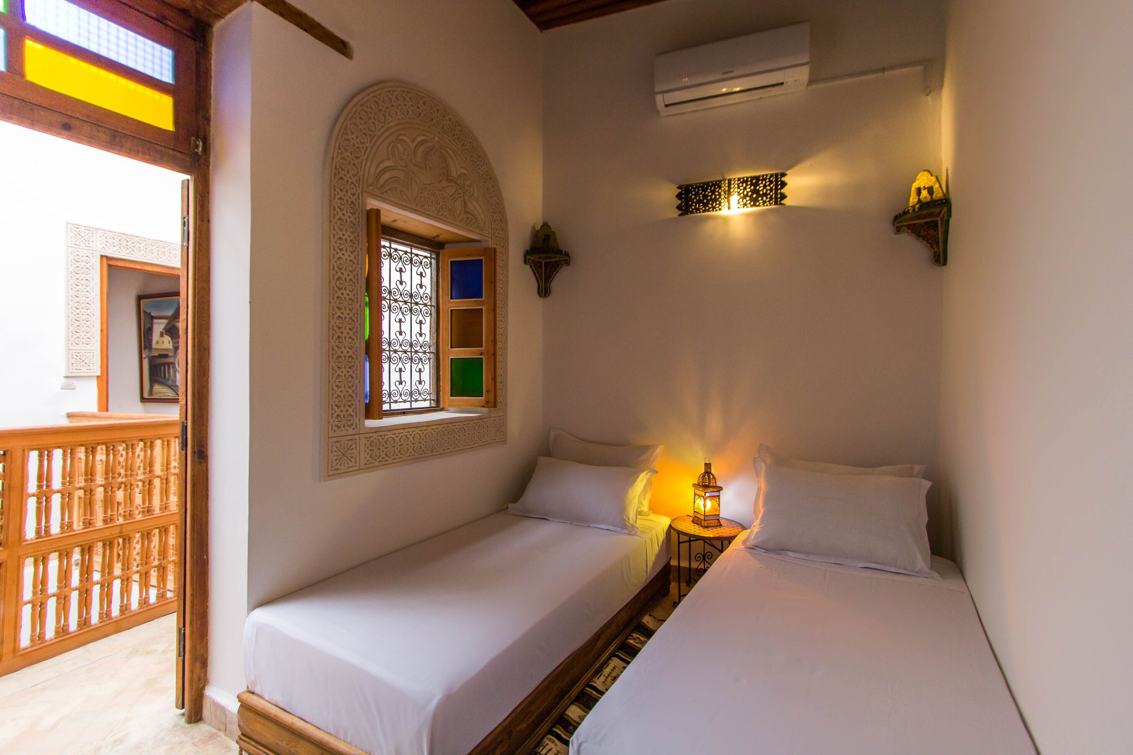 A room with two beds, a window with decorative glass, an air conditioning unit, and a small table with a lantern.