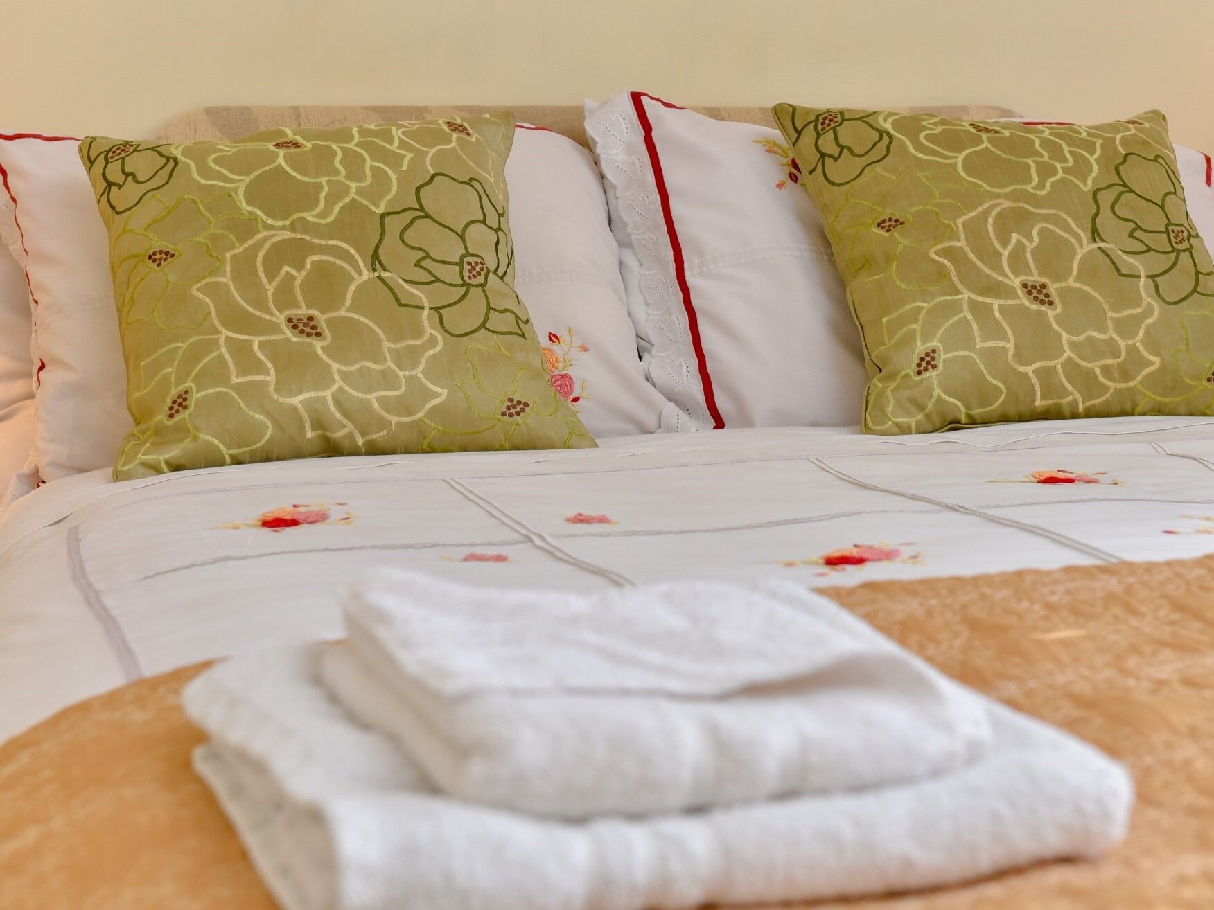 A neatly made bed with floral bedding, two green pillows with a floral pattern, and a stack of white towels.