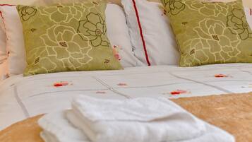 A neatly made bed with floral bedding, two green pillows with a floral pattern, and a stack of white towels.