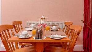 A dining table set with white teacups, saucers, a teapot, a sugar bowl, a creamer, a knife, a fork, a spoon, a glass of orange juice, and a vase with pink flowers. - The Warren Guest House (Carlisle)