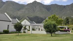 A modern building with a flat roof and white exterior walls, surrounded by a well-maintained lawn and a mountain backdrop. - Gaikou Lodge (Swellendam)