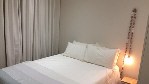 Una cama con ropa de cama blanca, una lámpara de noche y una cortina.