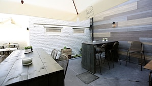 An outdoor dining area with a rustic wooden table and chairs, a white stone wall, and a wooden panel wall with a hanging sign.
