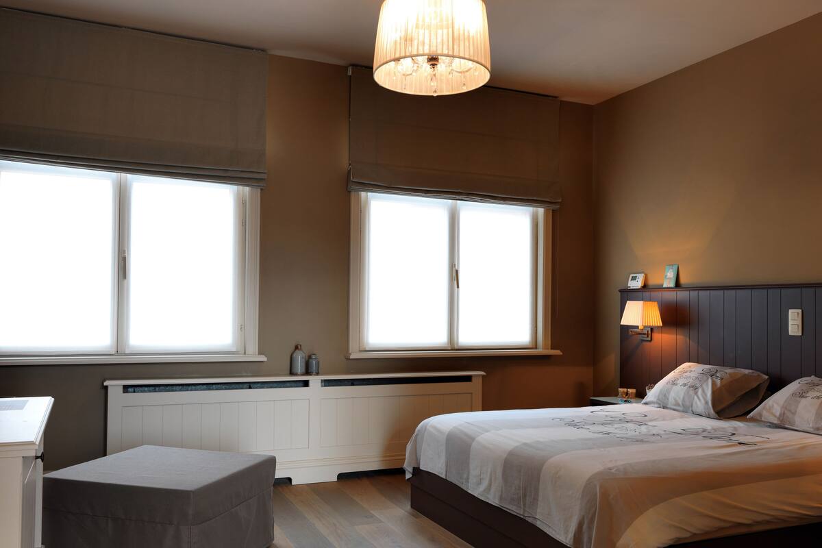 Quarto com cama, mesa de cabeceira, abajur e janelas amplas.