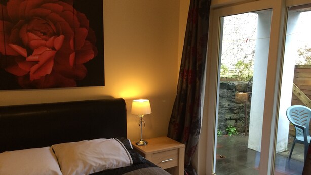 A bedroom with a bed, a nightstand with a lamp, a large window with curtains, and a view of the outside. - Glengorm Guest House (Oban)