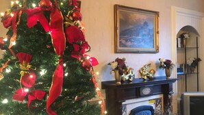 A living room with a Christmas tree decorated with lights and ornaments, a fireplace, a painting on the wall, and a television. - Glengorm Guest House (Oban)