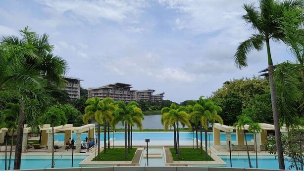 A resort pool area with palm trees, a clear sky, and a view of buildings in the distance. - Dr Calayan's 2BR Condo at Pico De Loro (Nasugbu)