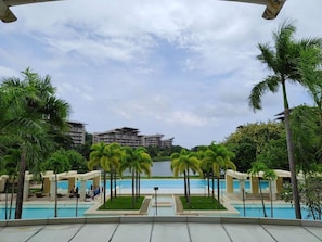 A resort pool area with palm trees, a clear sky, and a view of buildings in the distance. - Dr Calayan's 2BR Condo at Pico De Loro (Nasugbu)