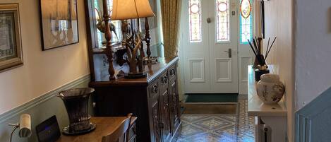 A hallway with a wooden table, chairs, a lamp, and a decorative mirror. There is a door with stained glass windows and a patterned floor.