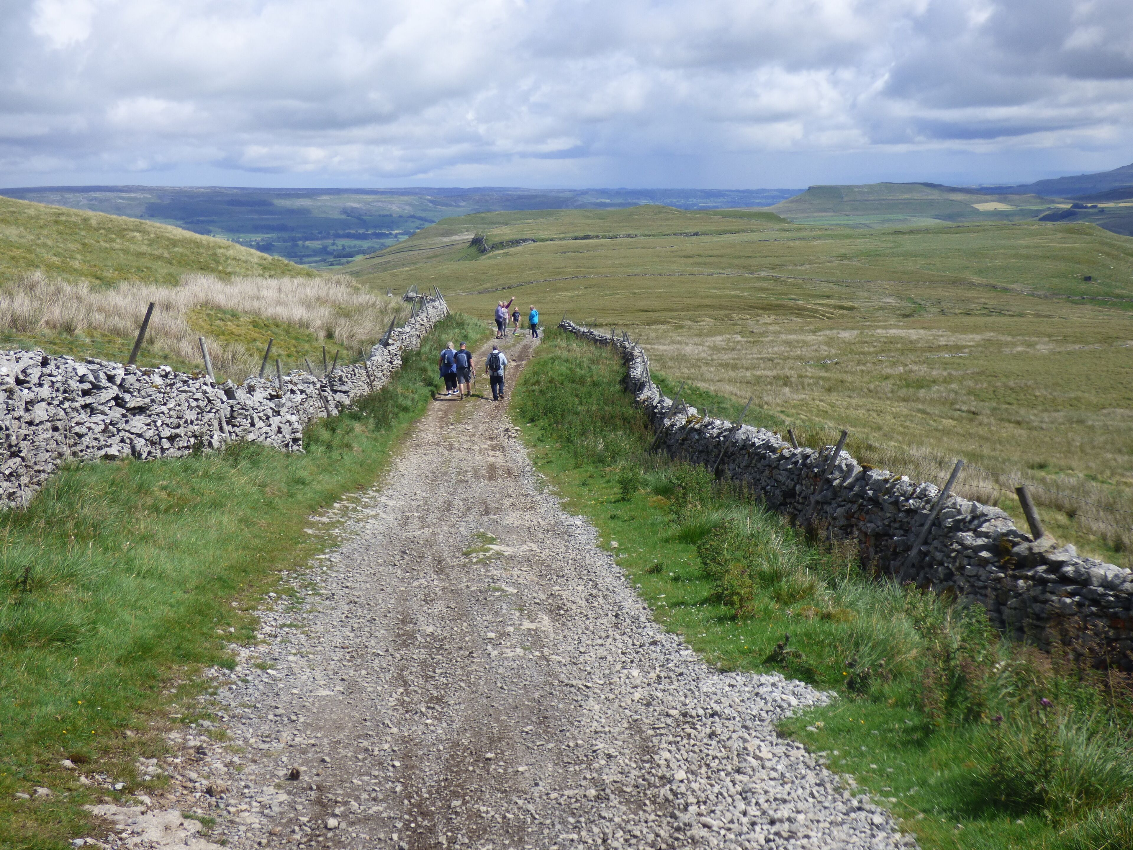 Ein Kiesweg, der von Steinmauern gesäumt wird und durch eine grüne Landschaft zu weiter Hügeln führt.