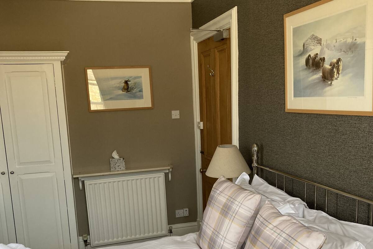 Quarto de hotel com cama, radiador, porta, quadro emoldurado e luminária.
