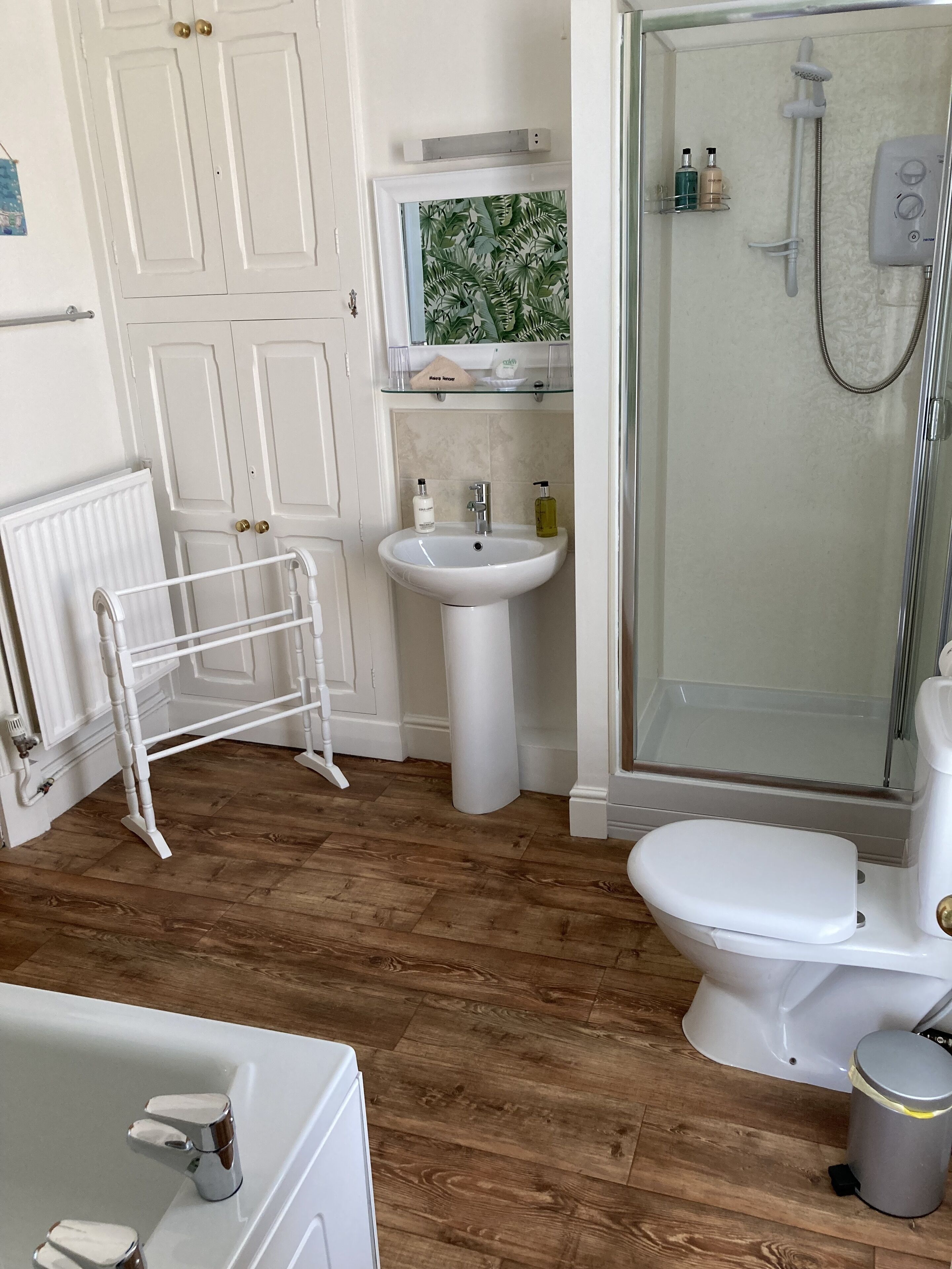 A bathroom with a white sink, toilet, and shower. There is a radiator, a towel rack, and a shelf with toiletries.