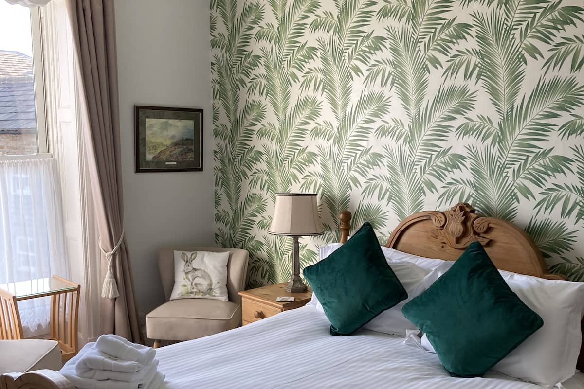 Cama bem arrumada com lençóis brancos e duas almofadas verdes, cabeceira de madeira e mesa de cabeceira com abajur.