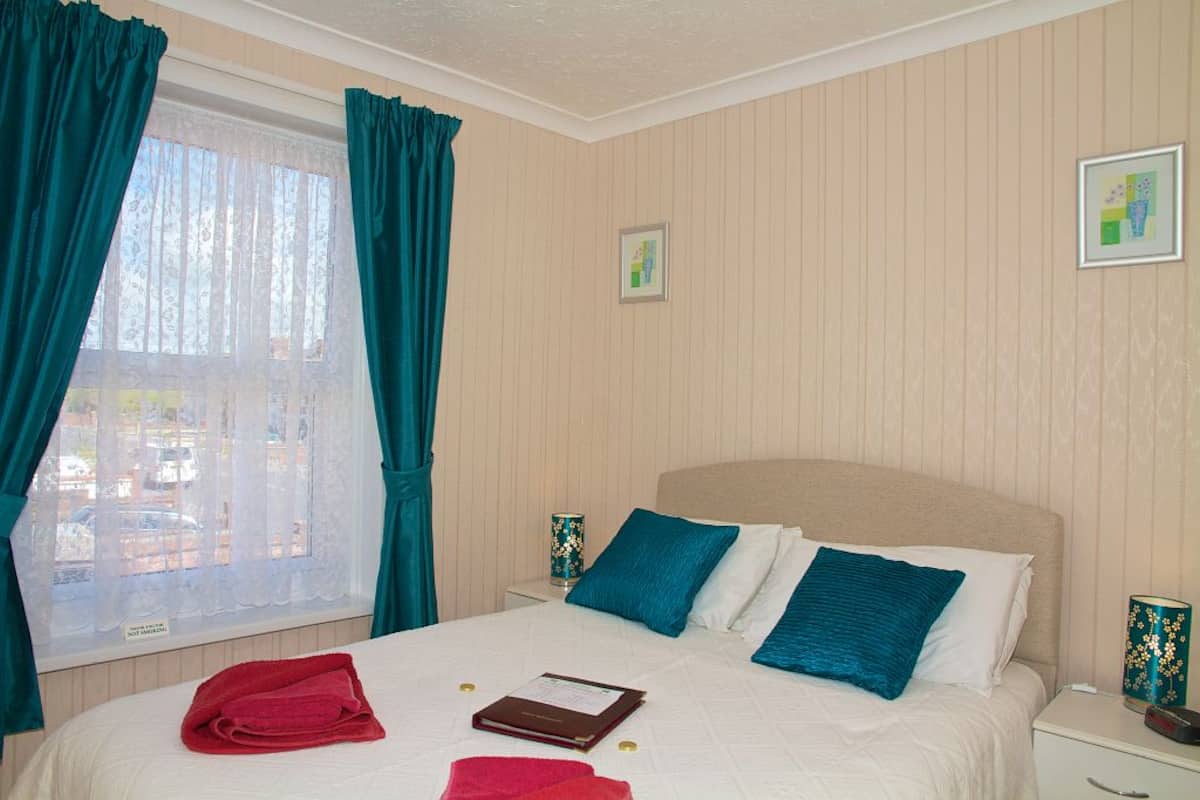 A neatly made bed with blue and white pillows, a red towel, and a small brown book on the bed. There is a window with teal curtains, a framed picture on the wall, and a nightstand with a decorative lamp.