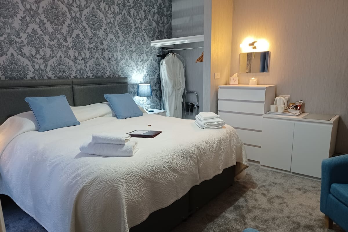 A neatly made bed with a white comforter and two pillows, a bedside table with a lamp, a mirror, a small cabinet, and a blue chair.