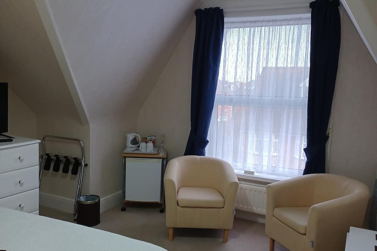 A bedroom with a bed, two armchairs, a small table, a radiator, and a window with curtains.
