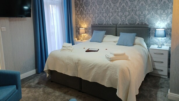 A neatly made bed with white linens, a bedside lamp, and a nightstand with a lamp. - Palm Court (Weymouth)