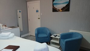 A hotel room with a bed, two blue armchairs, a small table with a phone, a painting of a beach scene, and a white door. - Palm Court (Weymouth)