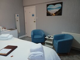Una habitación de hotel con una cama, dos sillones azules, una mesita con un teléfono, un cuadro de una playa y una puerta blanca.