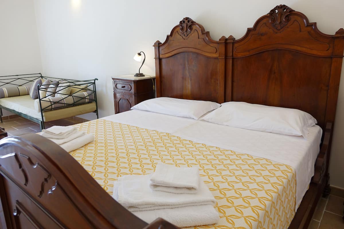A neatly made bed with a patterned bedspread and pillows, a wooden nightstand with a lamp, and a metal bench with cushions.