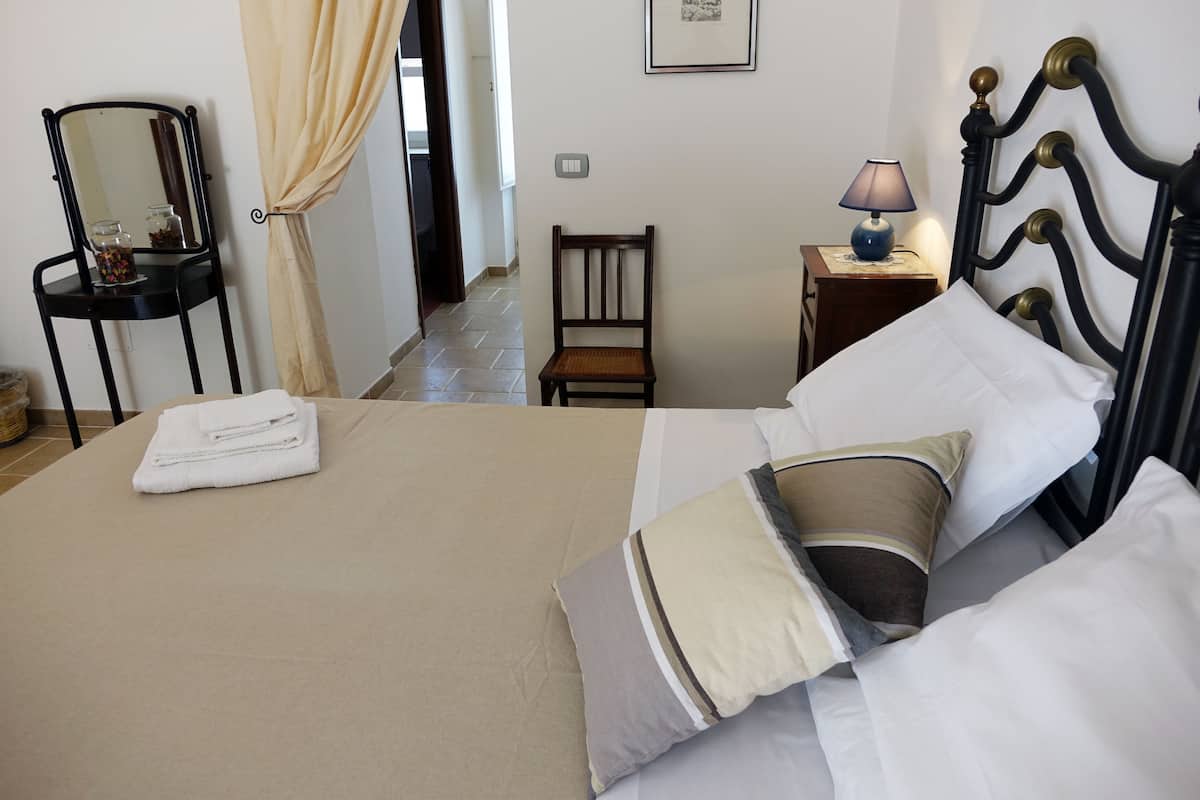 A neatly made bed with a beige bedspread, two pillows, and a folded white towel. There is a small wooden chair, a nightstand with a lamp, and a framed picture on the wall.