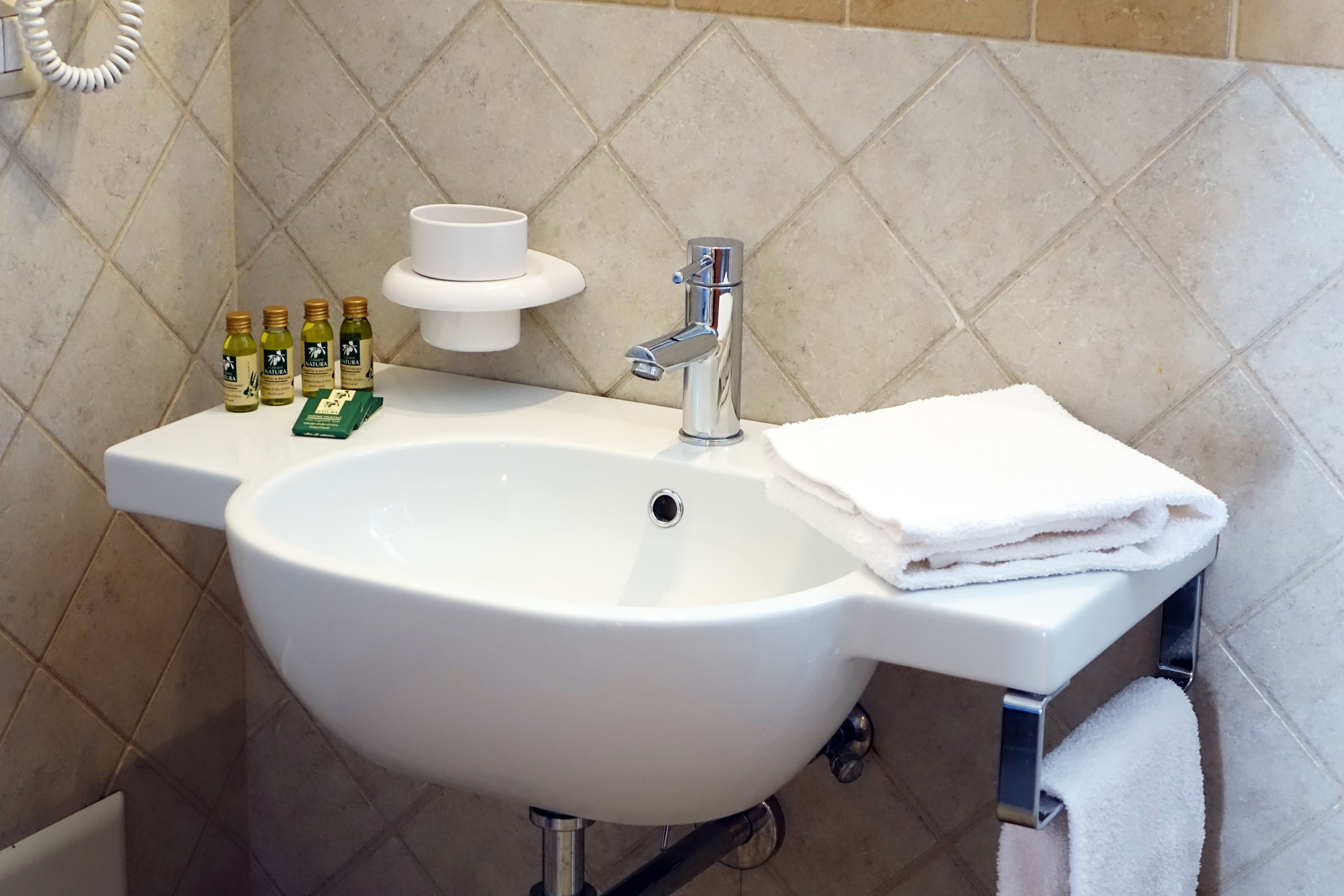 Un lavabo de salle de bain avec un bac blanc, un robinet chromé et un porte-serviettes sur lequel est posée une serviette pliée. On peut voir quatre bouteilles d’huile d’olive et un distributeur de savon sur le bord du lavabo.