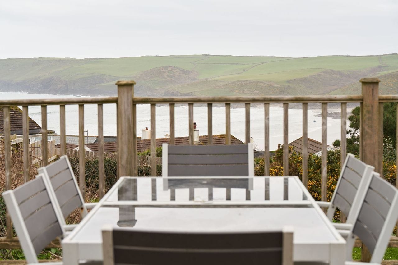 A table and chairs on a balcony overlooking a coastal landscape with hills and a view of the sea.