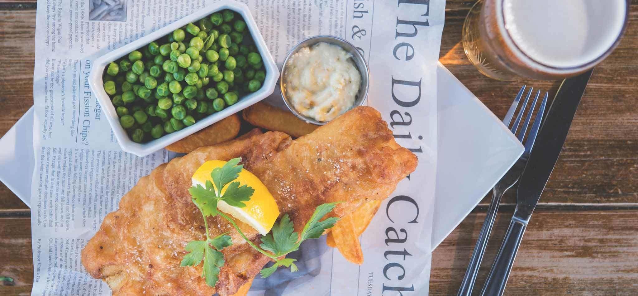 A meal of fish and chips, peas, and sauce on a newspaper.